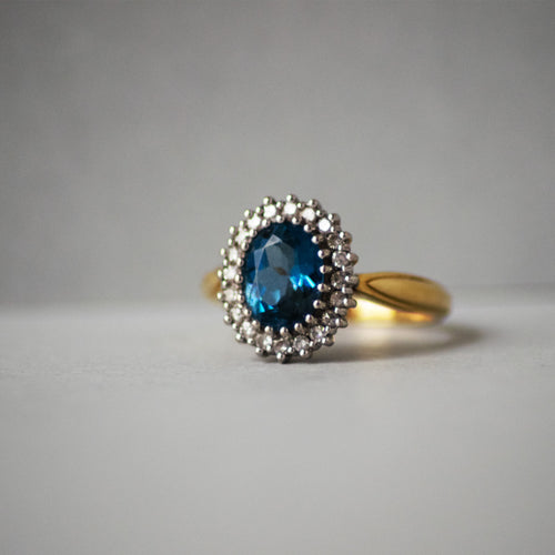 Gold ring with a blue gemstone and diamond-studded halo on a gray background